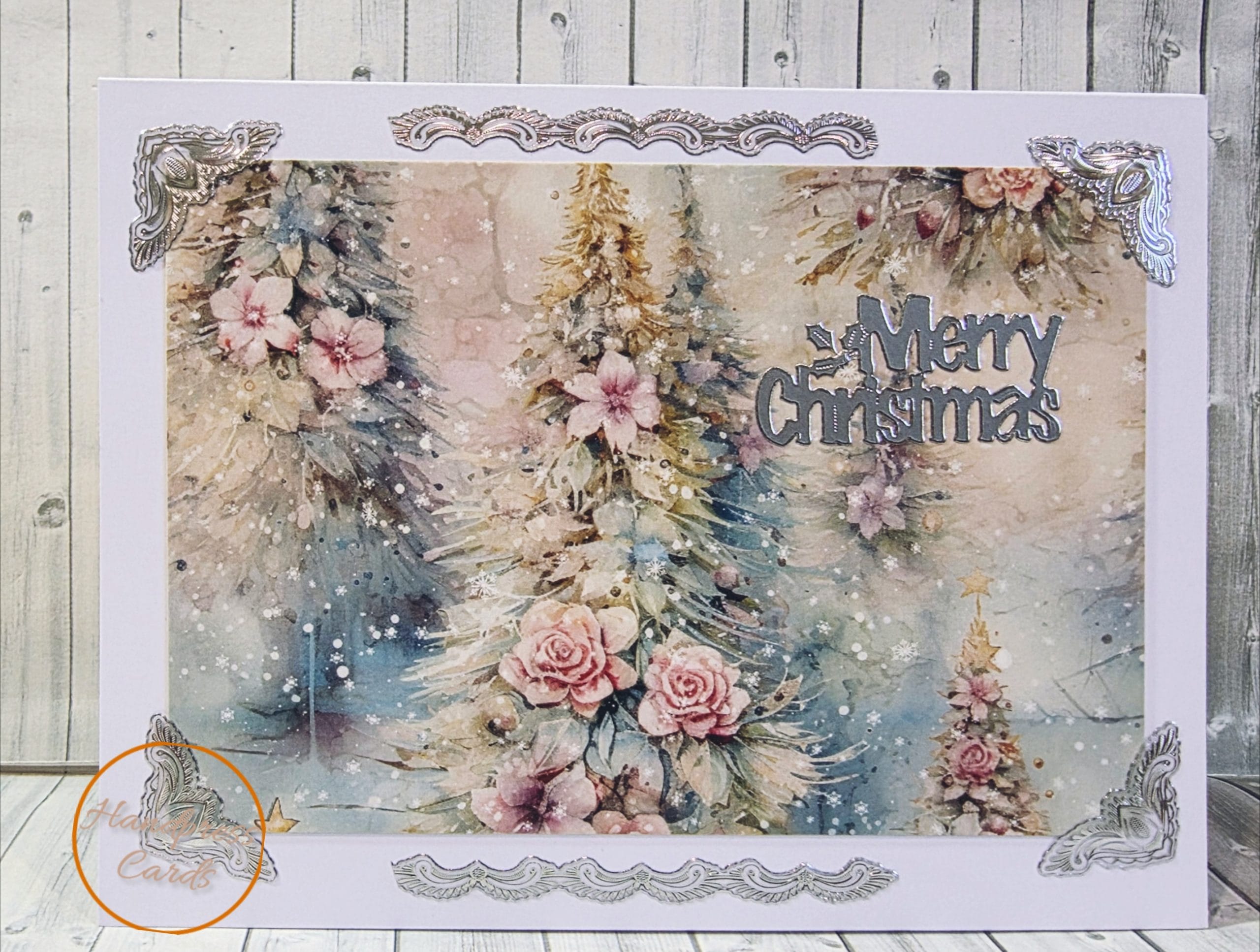 Handmade Christmas card with snowy trees and pink roses in soft watercolour, silver foil corners and borders and a silver Merry Christmas sentiment