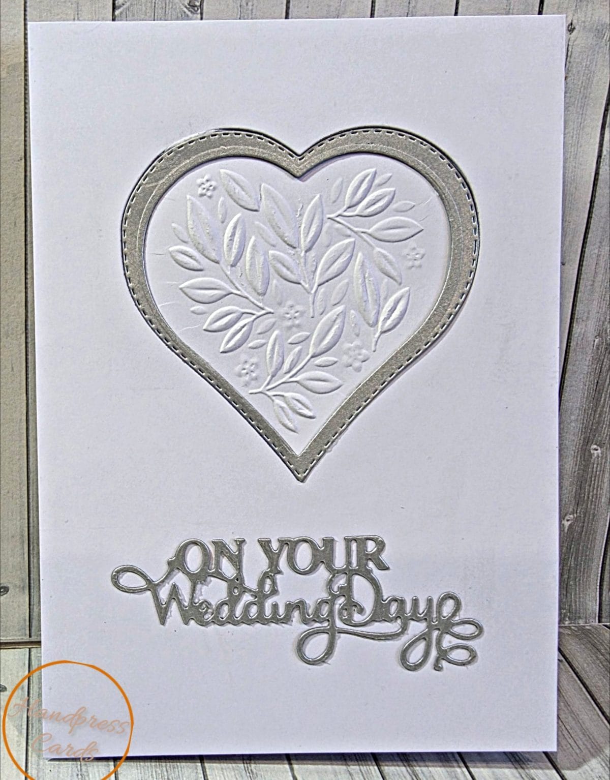 Handmade white wedding card with an embossed leaf and flower heart in a silver stitched frame and a silver On Your Wedding Day sentiment