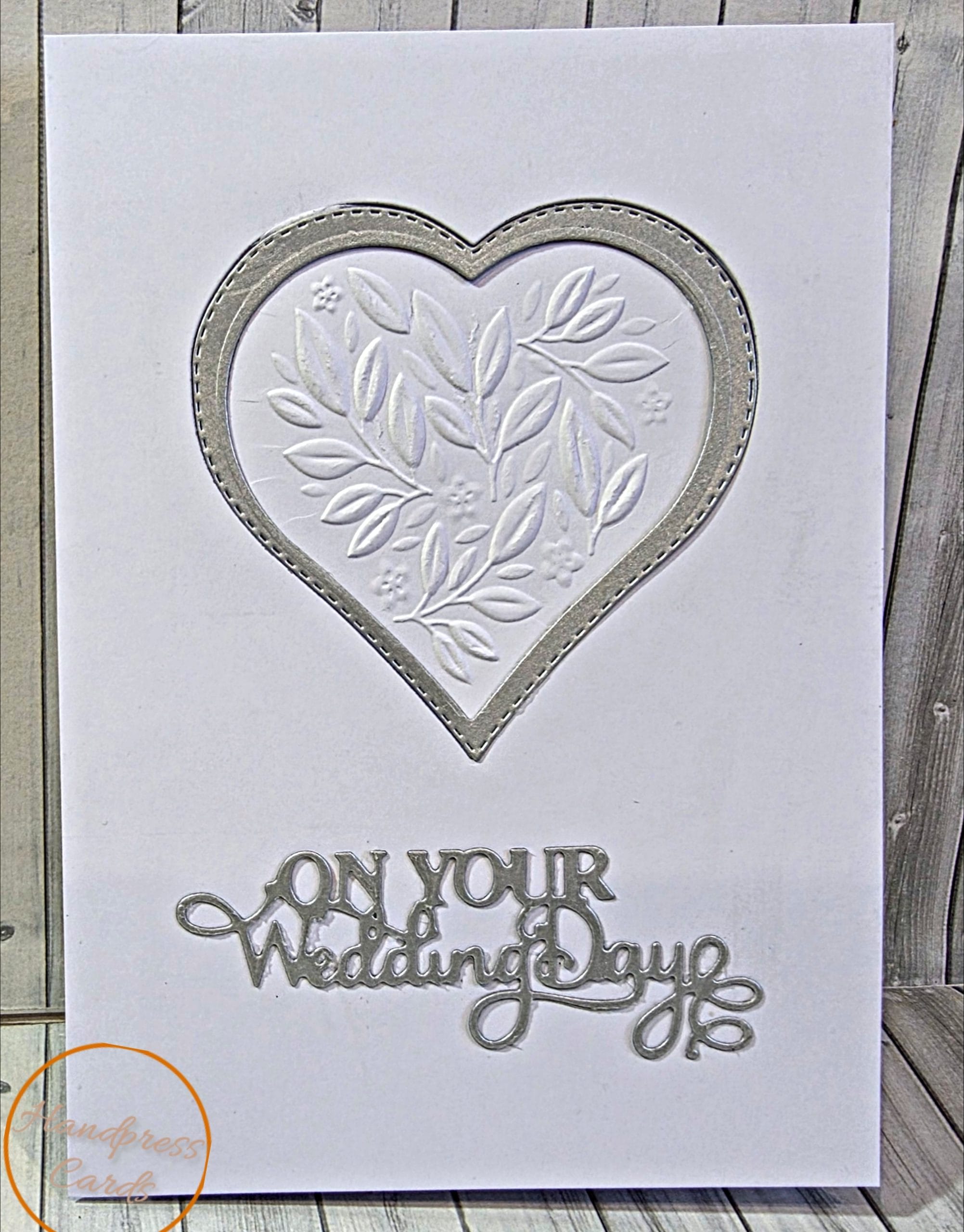 Handmade white wedding card with an embossed leaf and flower heart in a silver stitched frame and a silver On Your Wedding Day sentiment